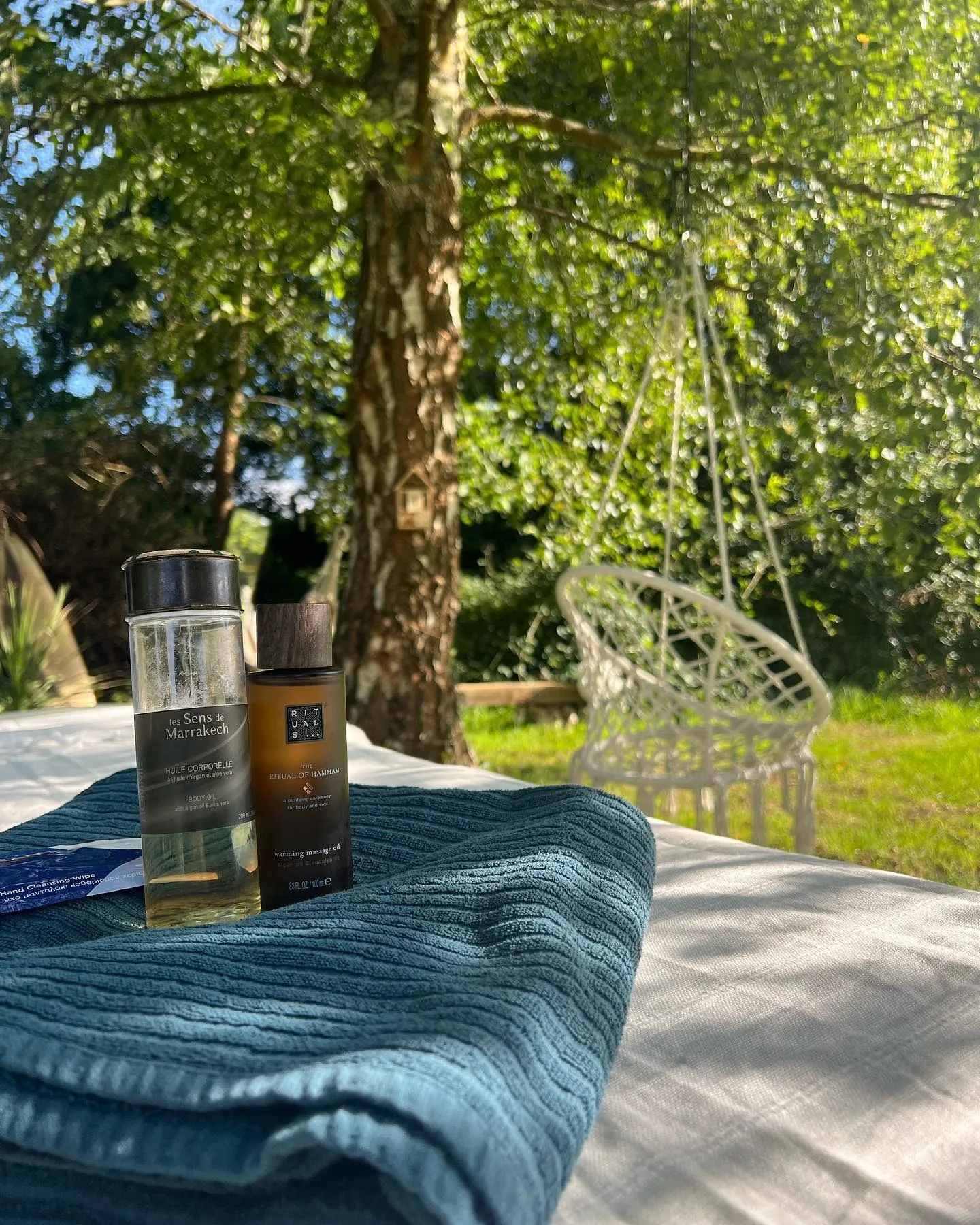 Huiles de massage Rituals posées sur la table, serviette bleue, jardin verdoyant en arrière-plan
